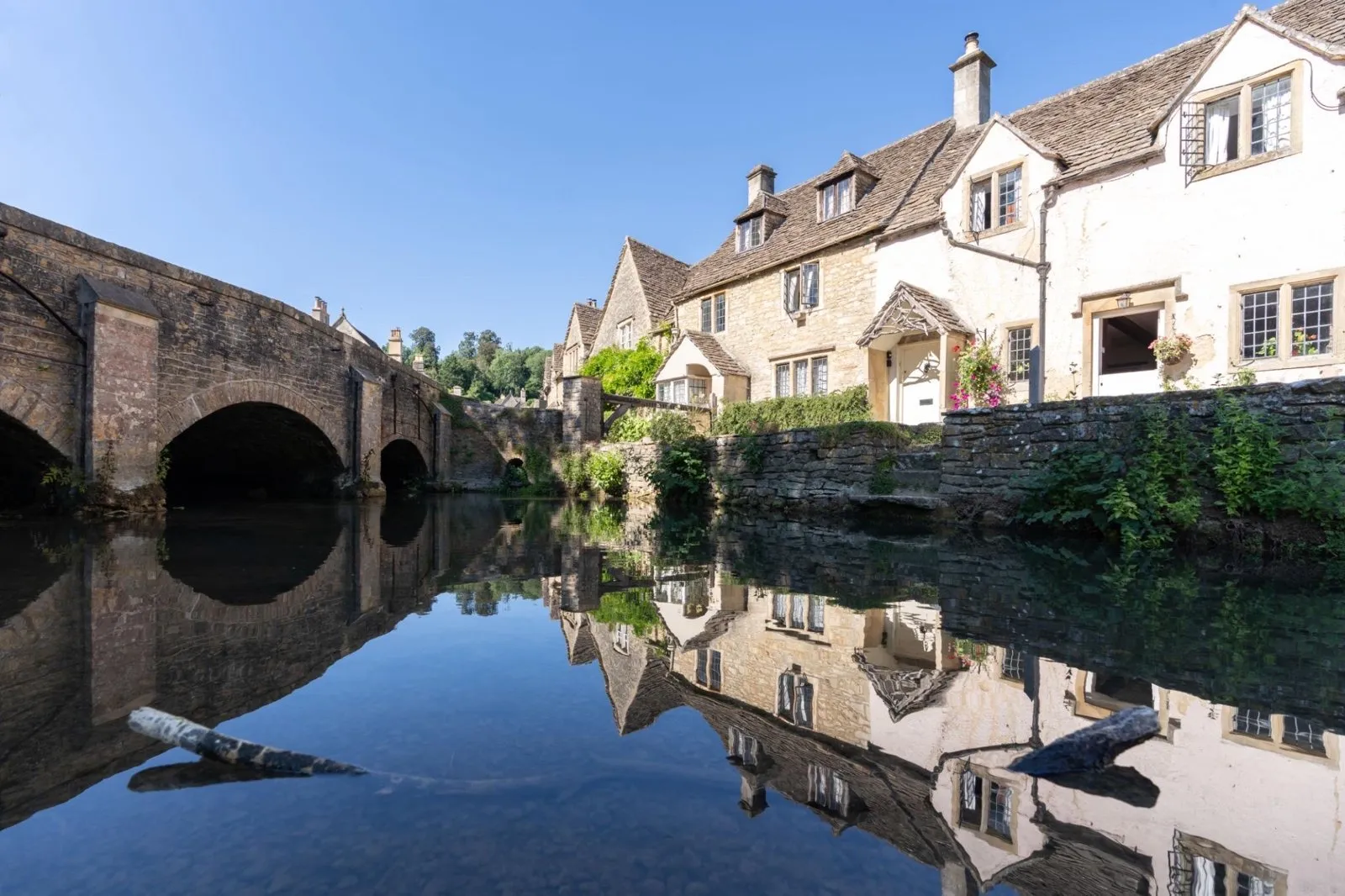 Cotswolds stone cottages reflected in a calm river next to an arched bridge.