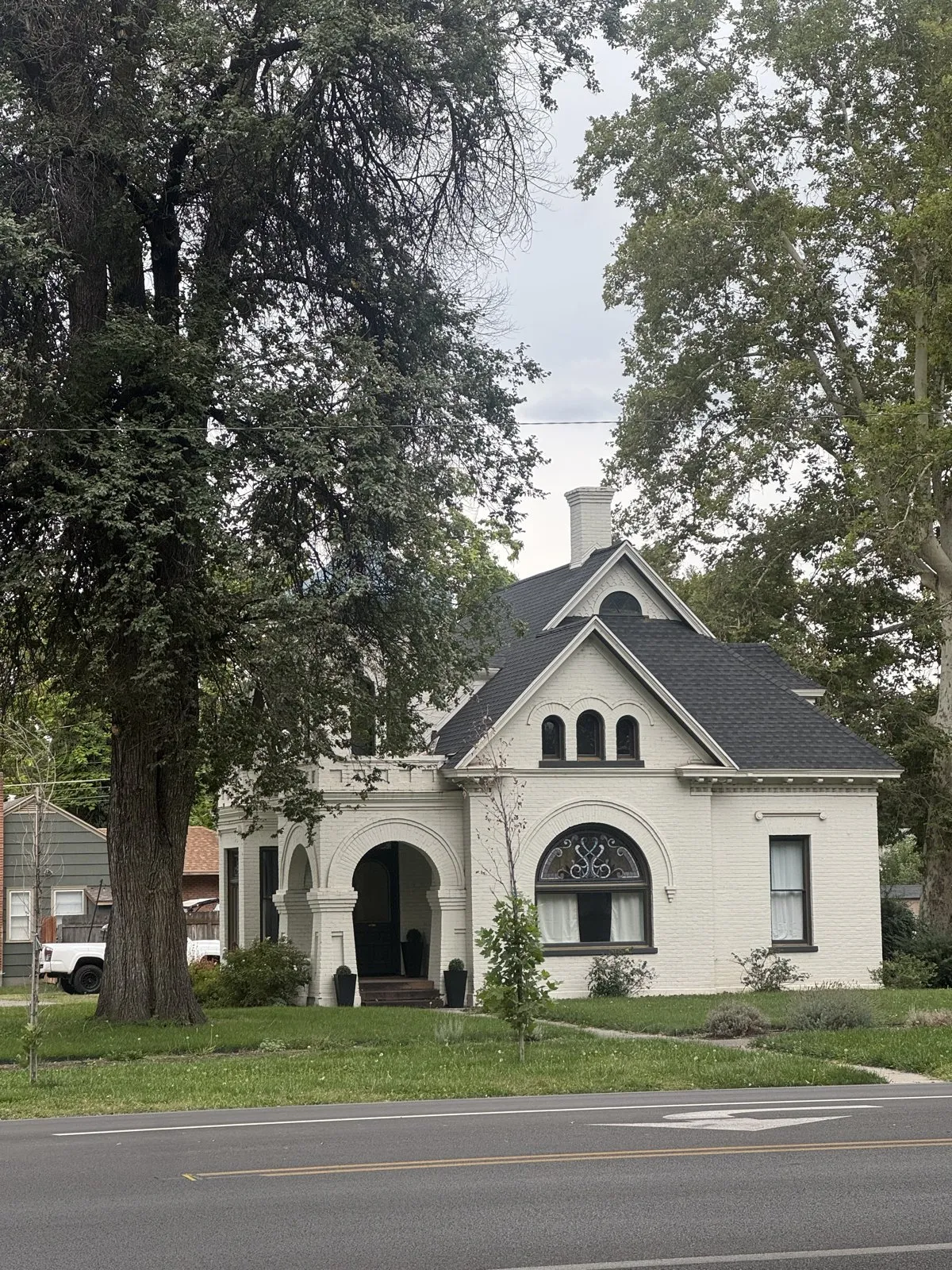 A white brick historic home with arched windows and a black roof alongside a paved street.