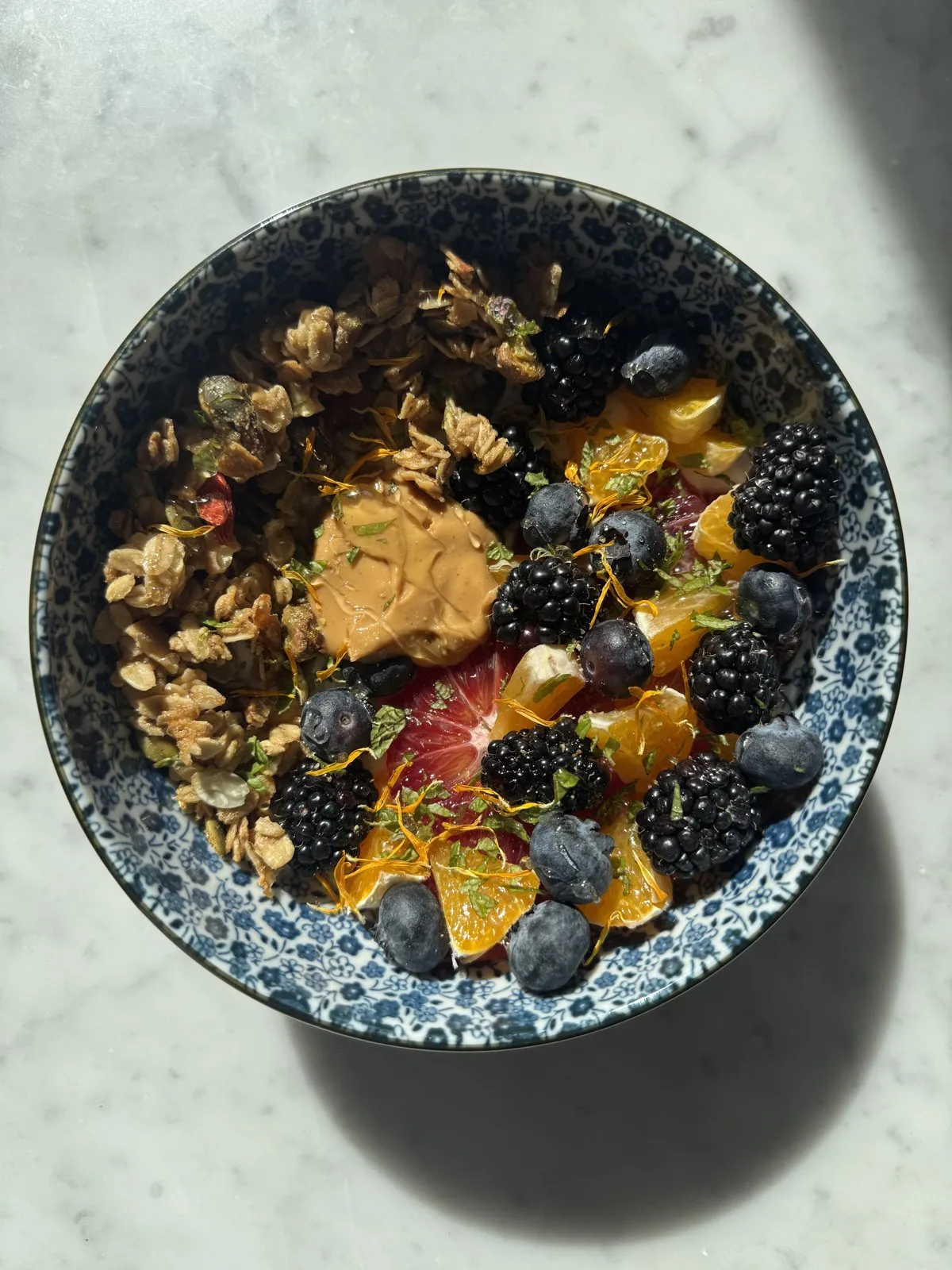 A patterned ceramic bowl filled with yogurt, sliced blood oranges, mandarins, blackberries, blueberries, granola, and peanut butter.