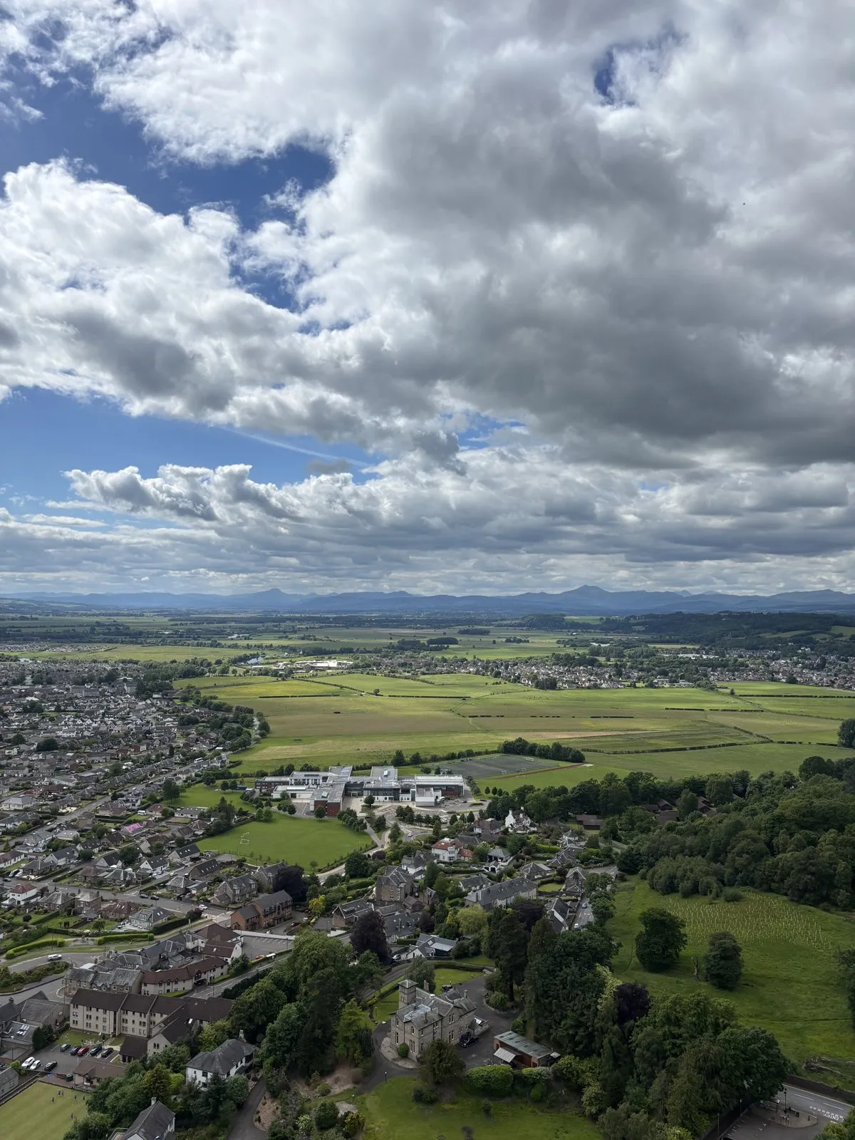 High aerial view of a town, green fields, and distant mountains beneath puffy clouds.