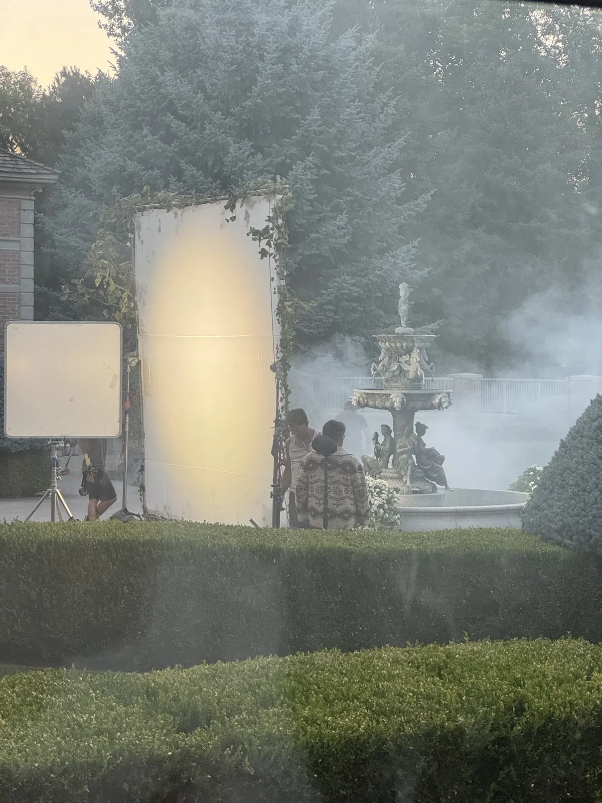 A film production set featuring large light diffusion panels and artificial smoke around an ornate fountain.